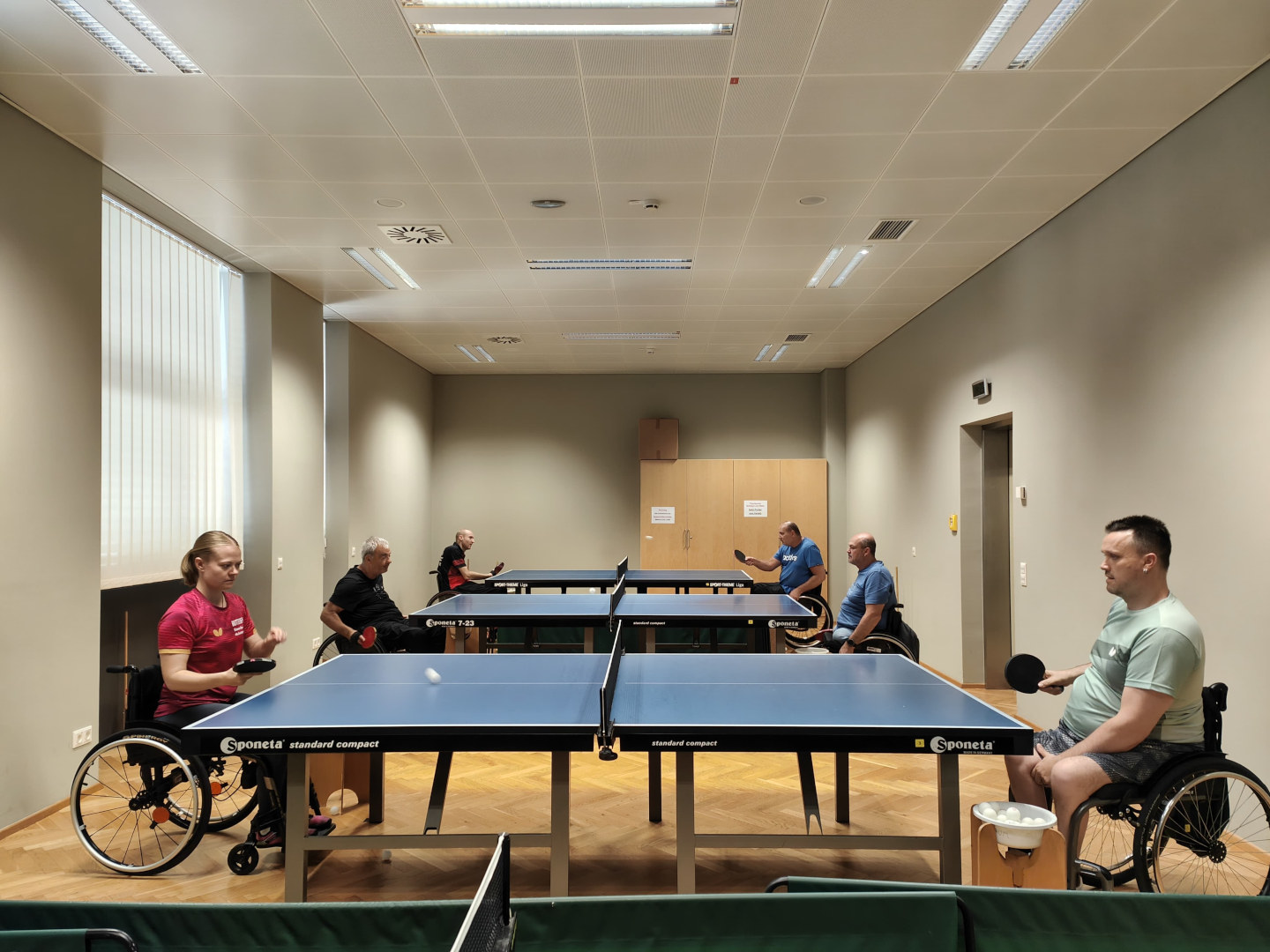 Rollstuhltischtennis: Im Bild sind eine Spielerin und fünf Spieler an drei Platten