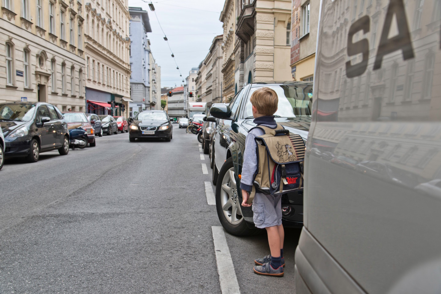 Schulbub blickt zwischen parkenden Fahrzeugen auf einen herannahenden PKW: