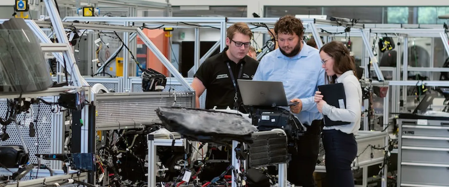 Zwei junge Männer und eine junge Frau schauen in einer Produktionshalle gemeinsam auf einen Laptop. Zwei junge Männer und eine junge Frau schauen in einer Produktionshalle gemeinsam auf einen Laptop.