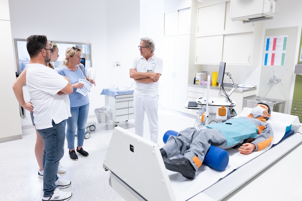 Ein Mitarbeiter des RH Häring spricht mit drei Besucher:innen in einem Behandlungsraum, auf der Liege liegt ein Patient, bei dem es sich jedoch zu Demonstrationszwecken um eine Puppe handelt