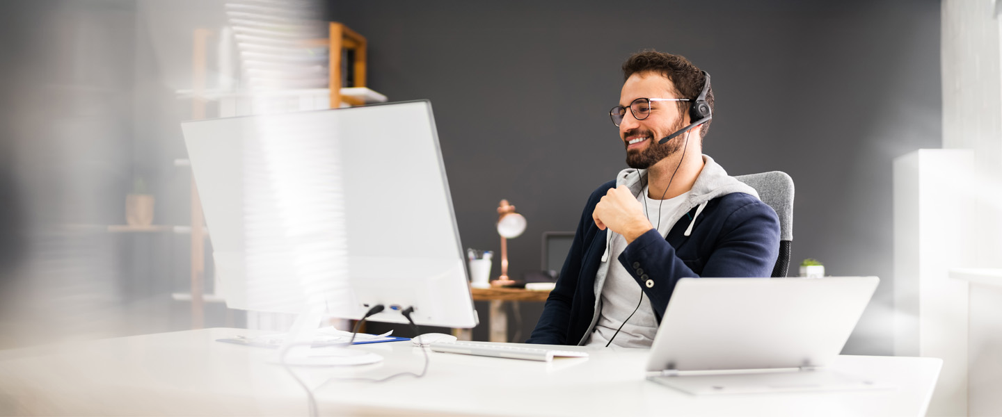 Ein Mann mit Headset sitzt an einem Schreibtisch und schaut freundlich lächelnd auf einen Bildschirm.