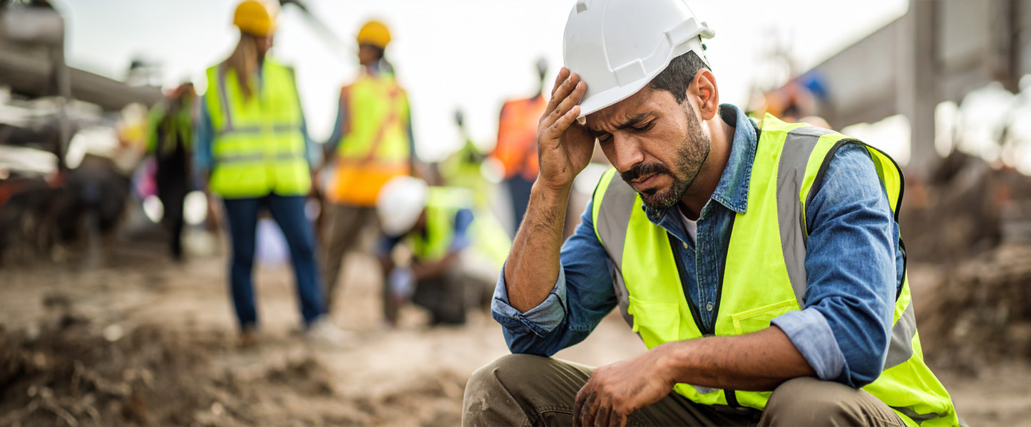 Erschöpft wirkender Bauarbeiter sitzt auf Baustelle und stützt Kopf in die Hand. Im Hintergrund sind andere Bauarbeiter zu sehen.
