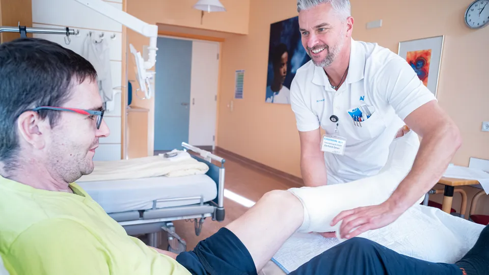 Ein Arzt behandelt in einem Krankenhauszimmer einen im Bett liegenden Patienten, der am linken Unterschenkel und Fuß einen Verband trägt. Ein Arzt behandelt in einem Krankenhauszimmer einen im Bett liegenden Patienten, der am linken Unterschenkel und Fuß einen Verband trägt.