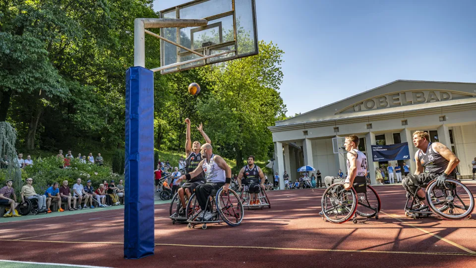 Mehrere Spieler mitten im Rollstuhlbasketballspiel, im Hintergrund Zuschauer