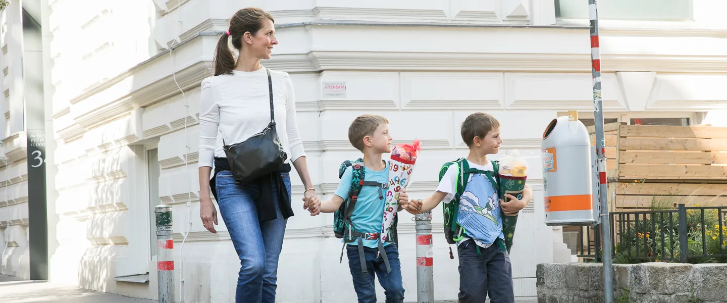 1.2.2_mein-sicherer-Schulweg_20210910-IMG_3291_web©MartinNußbaum.jpg Eine Mutter überquert mit ihren zwei Söhnen am Schutzweg eine Straße. Alle blicken nach links, um zu kontrollieren, dass kein Fahrzeug kommt.