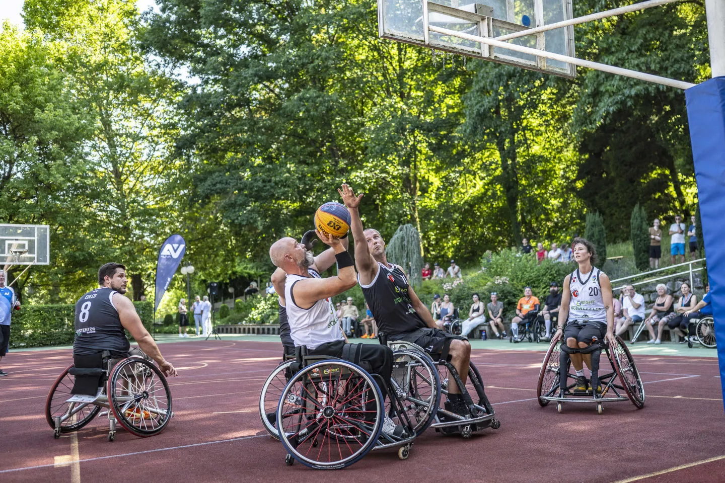 Mehrere Spieler mitten im Rollstuhlbasketballspiel, im Hintergrund Zuschauer
