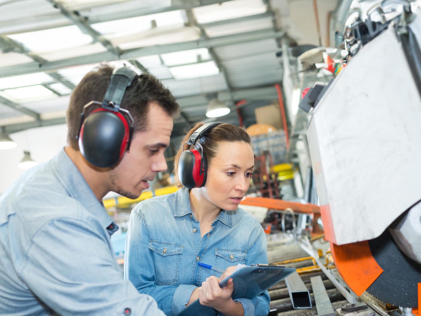 Ein Mann und eine Frau mit Kapselgehörschutz inspizieren eine Maschine in einer Produktionshalle.