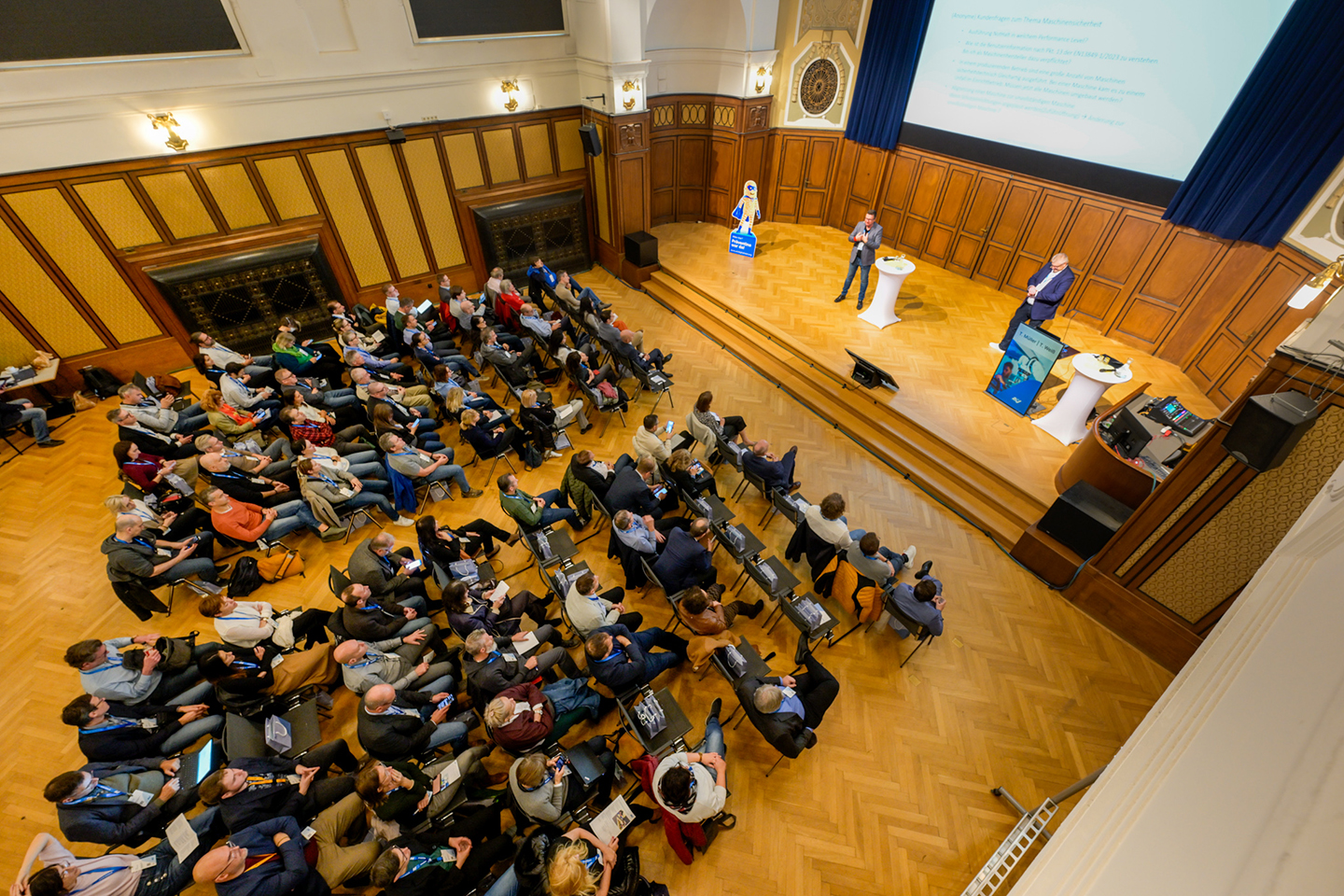 Blick von oben in einen Vortragssaal. Viele Menschen sitzen im Publikum, zwei stehen auf der Bühne
