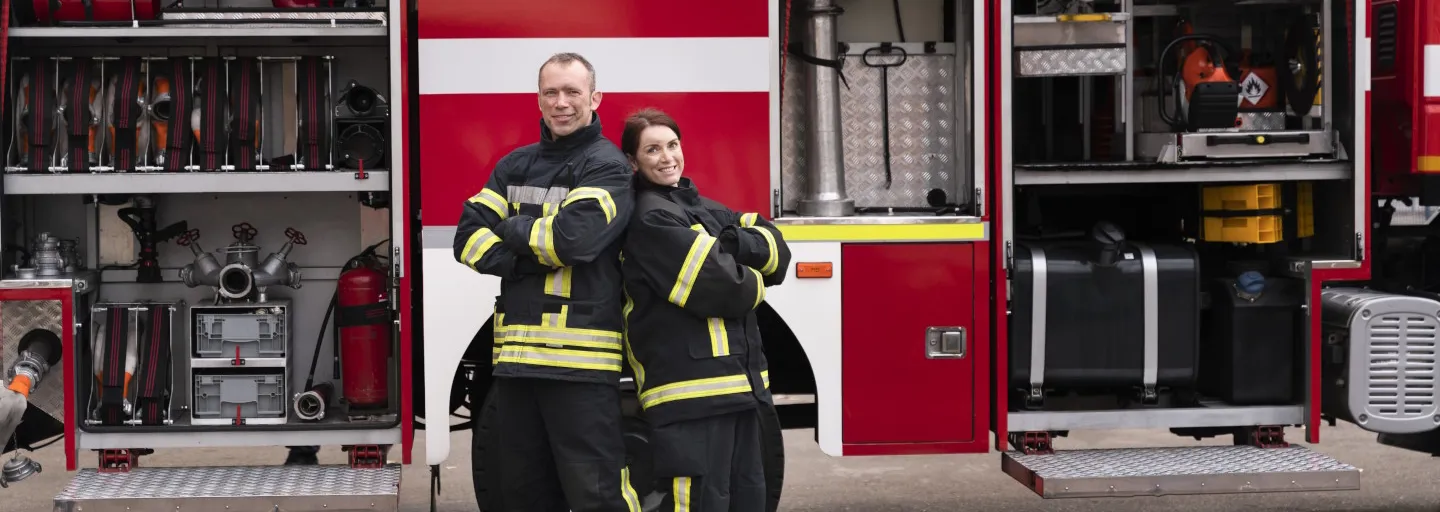 weißer Feuerwehr Schriftzug auf rotem Feuerwehrauto Feuerwehrmann und Feuerwehrfrau stehen nebeneinander vor einem Feuerwehrfahrzeug