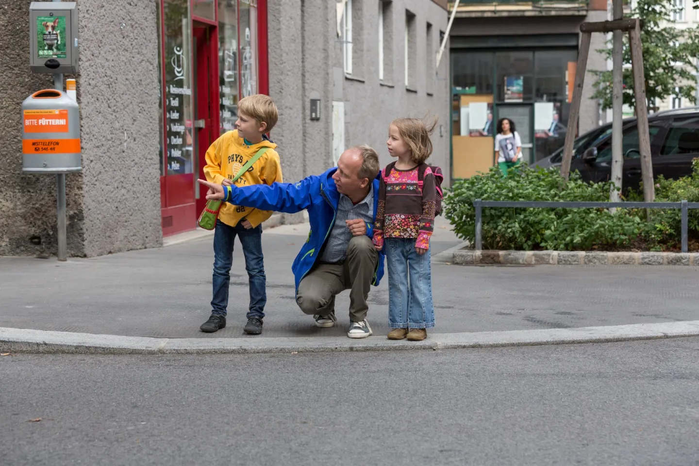 Schulweg 1440 Foto P.Winkler AUVA Mann hockt zwischen zwei Kindern am Gehsteigrand und deutet auf die Straße