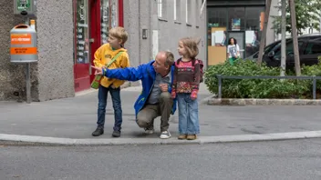 Schulweg 1440 Foto P.Winkler AUVA Mann hockt zwischen zwei Kindern am Gehsteigrand und deutet auf die Straße