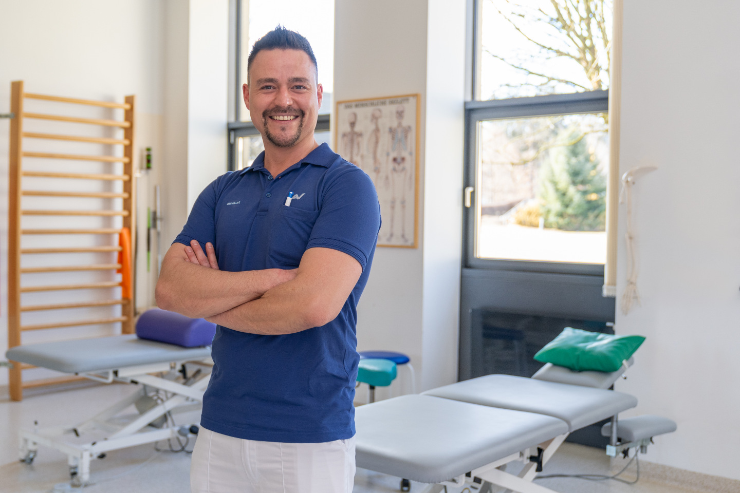 Ein Physiotherapeut steht mit verschränkten Armen vor zwei Liegen, im Hintergrund ist auch eine Sprossenwand