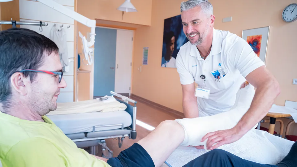 Ein Arzt behandelt in einem Krankenhauszimmer einen im Bett liegenden Patienten, der am linken Unterschenkel und Fuß einen Verband trägt.