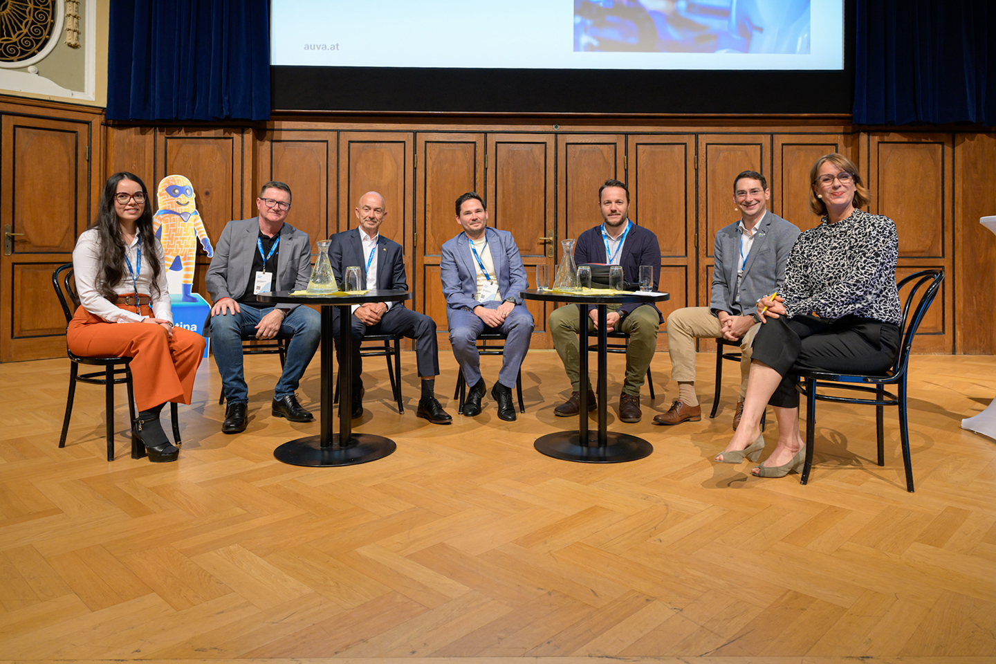 Mehrere Personen sitzen auf Sesseln in einem Halbkreis. Vor ihnen stehen zwei runde Tische