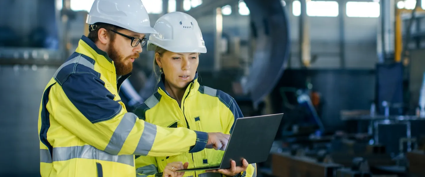 Ein Mann und eine Frau mit Bauhelm und Warnwesten bekleidet schauen in einer Fabrikshalle auf einen Laptop