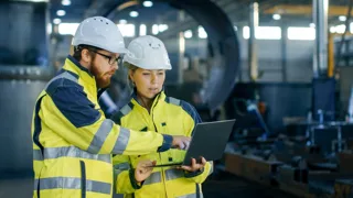 Ein Mann und eine Frau mit Bauhelm und Warnwesten bekleidet schauen in einer Fabrikshalle auf einen Laptop