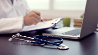 Ein Stethoskop liegt auf dem Schreibtisch vor einem nur halb sichtbaren Arzt, der am Laptop arbeitet Ein Stethoskop liegt auf dem Schreibtisch vor einem nur halb sichtbaren Arzt, der am Laptop arbeitet