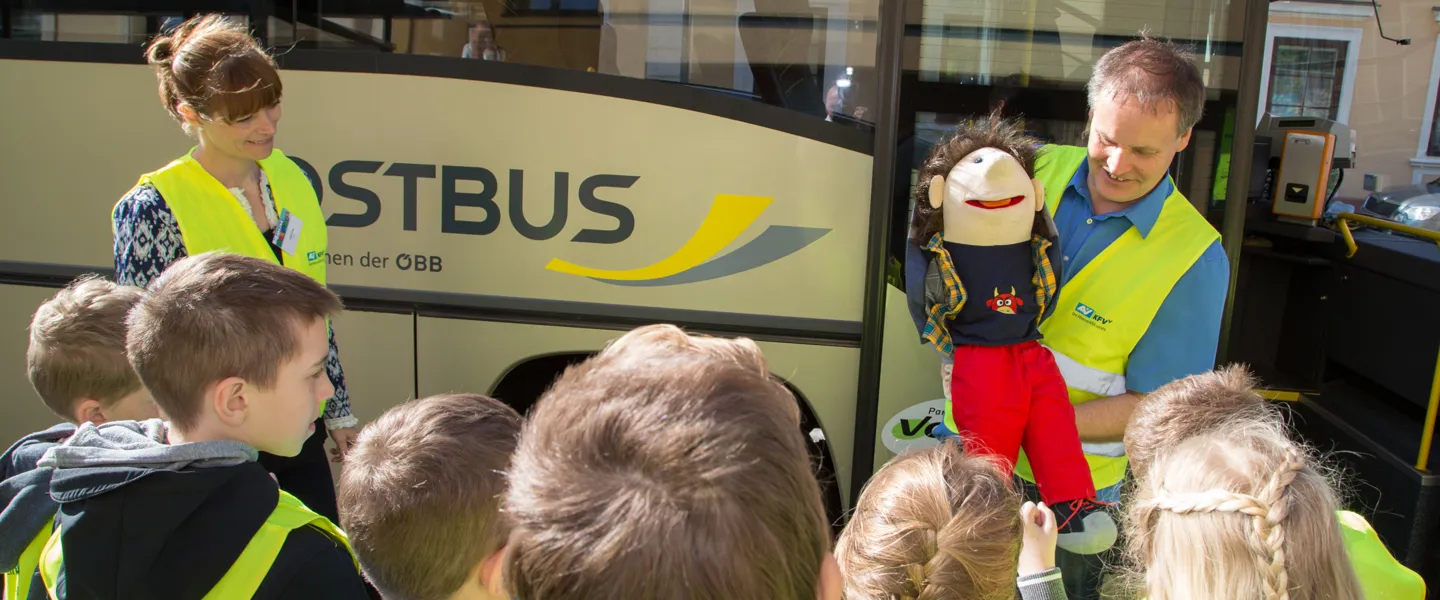 Mann mit Handpuppe spricht vor einem Bus zu einer Gruppe von Kindern
