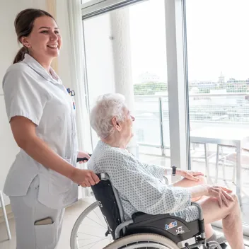 Pflege - Patient im Rollstuhl bei Aussichtsfenster Patient wird im Rollstuhl zum Glasfenster mit Aussicht ins Grüne geführt