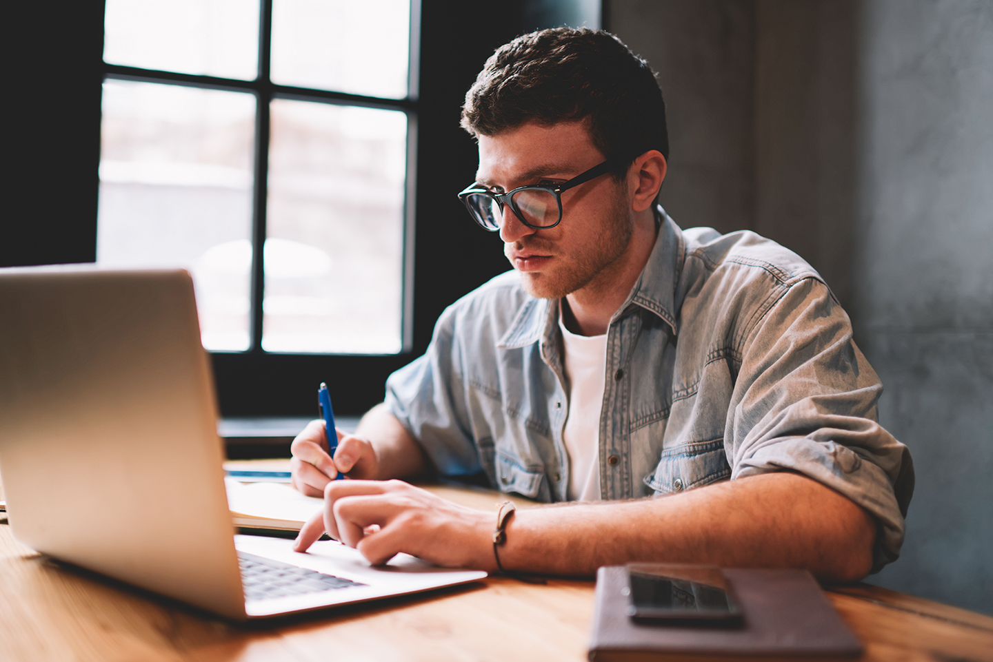 Ein Mann sitzt an einem Schreibtisch und schaut auf einen geöffneten Laptop. Mit der rechten Hand macht er Notizen auf einem Block.