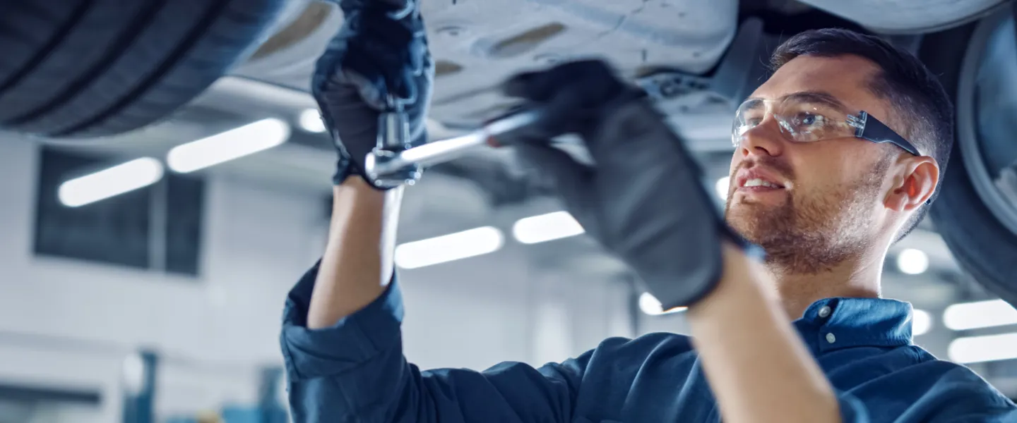 Mechaniker mit Schutzbrille und Schutzhandschuhen arbeitet in einer Werkstatt unter einem mit einer Hebebühne hochgefahrenen Auto