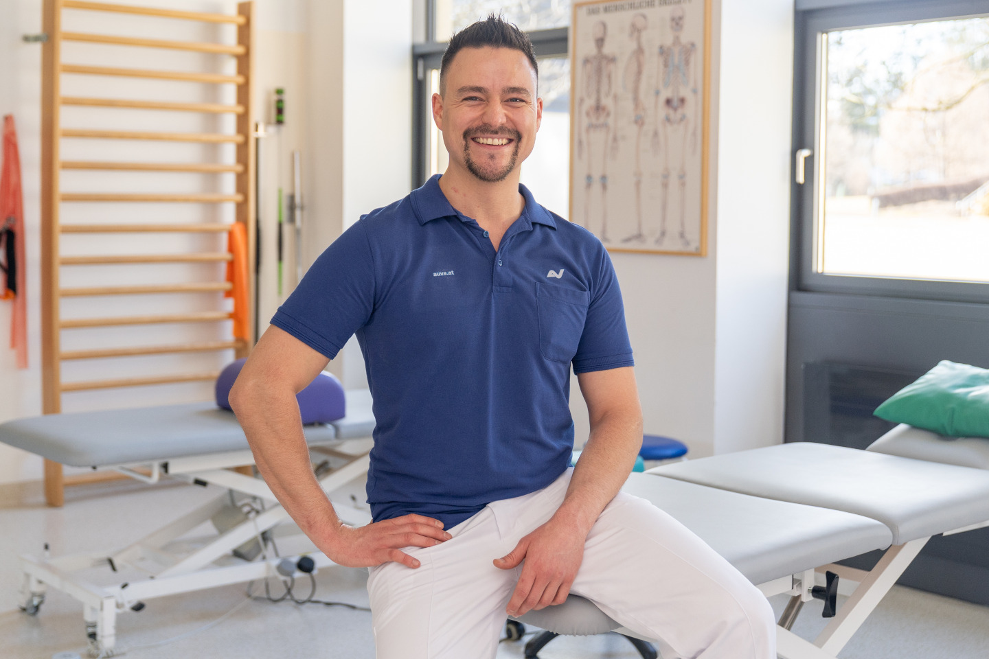 Ein Physiotherapeut sitzt auf einer Liege, im Hintergrund sind eine weitere Liege und eine Sprossenwand