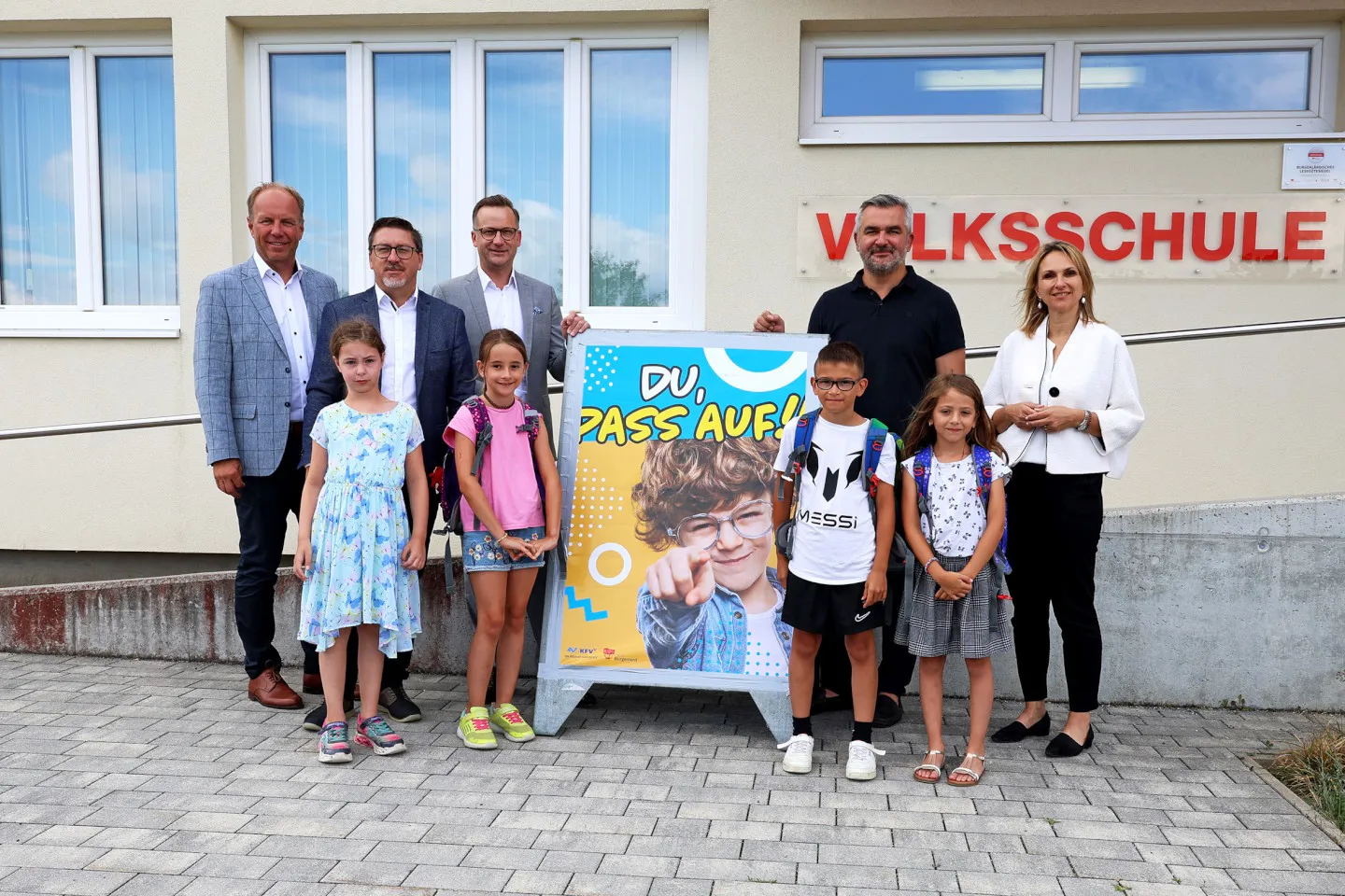 LR Dorner Schulstart Volksschule 001 1440 Credit Landesmedienservice Burgenland Erwachsene und Volksschulkinder mit Schulstart-Plakat vor der Volksschule