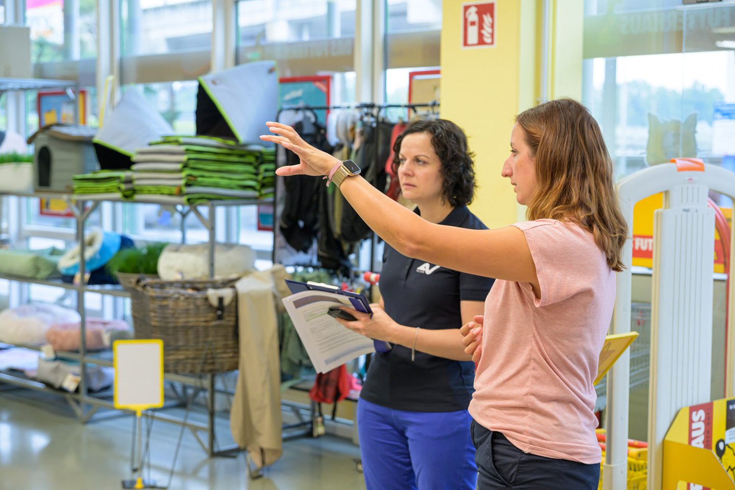 Zwei Frauen stehen in einem Geschäftslokal. Eine deutet mit dem linken Arm auf etwas, die andere blickt in die angedeutete Richtung.