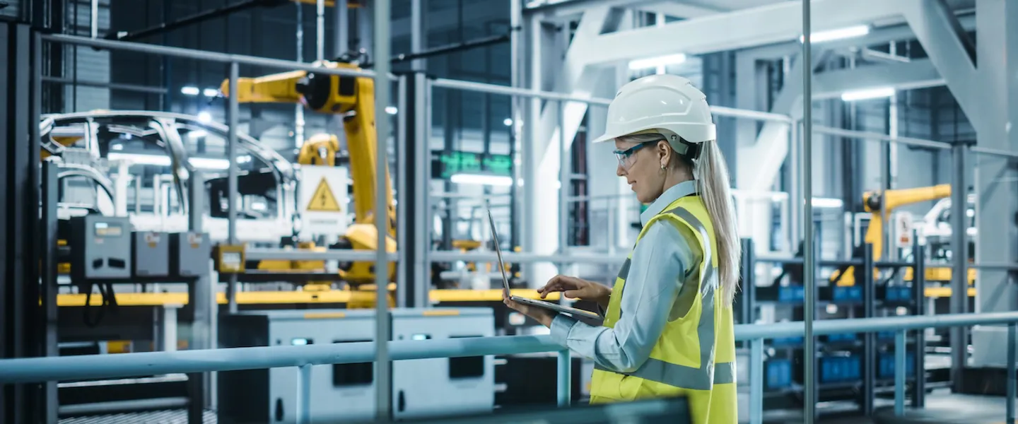Frau mit Helm in der Produktion einer Autofabrik steuert einen Roboterarm mit Laptop