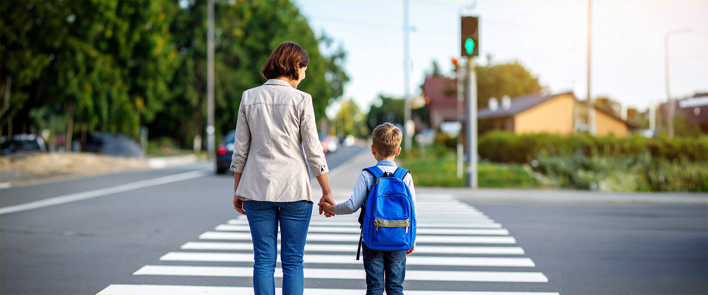 Rückansicht einer Frau und eines Kindes mit Schultasche. Sie stehen vor einem Zebrastreifen und blicken auf die gegenüberliegende grün leuchtende Fußgängerampel