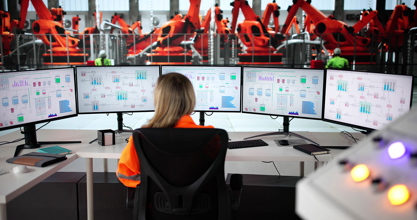 Eine Frau sitzt vor fünf Monitoren, die im Halbkreis vor ihr angeordnet sind. Im Hintergrund sieht man Industrieroboter