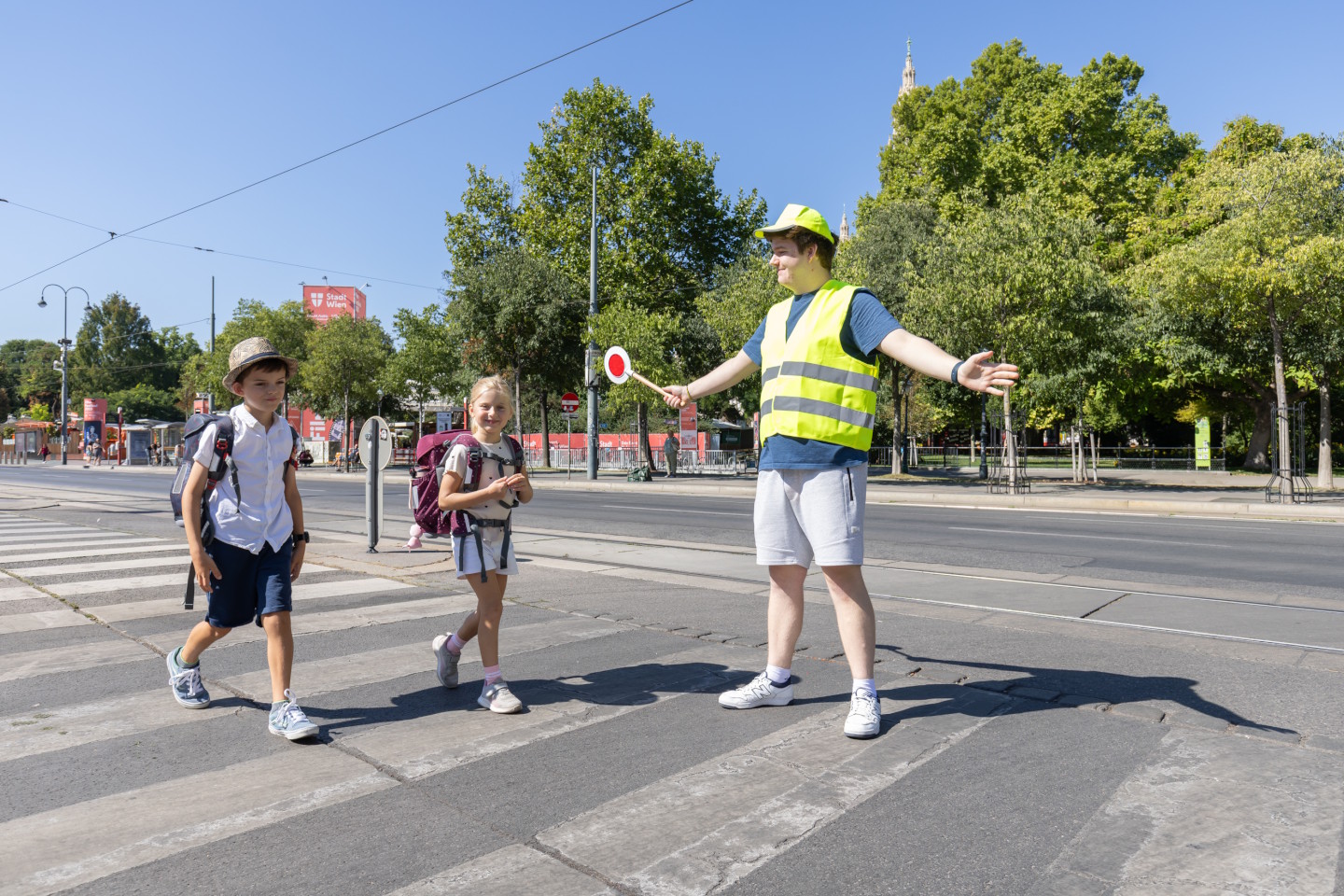 Schülerlotse mit Warnweste und Kelle und zwei Schulkinder am Zebrastreifen