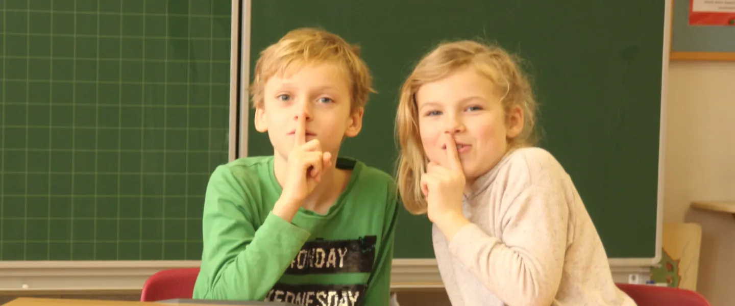 Zwei Kinder in einem Klassenraum machen mit ihrem Finger das Zeichen für "Still sein" Zwei Kinder in einem Klassenraum machen mit ihrem Finger das Zeichen für "Still sein"