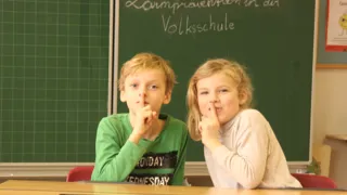 Zwei Kinder in einem Klassenraum machen mit ihrem Finger das Zeichen für "Still sein" Zwei Kinder in einem Klassenraum machen mit ihrem Finger das Zeichen für "Still sein"