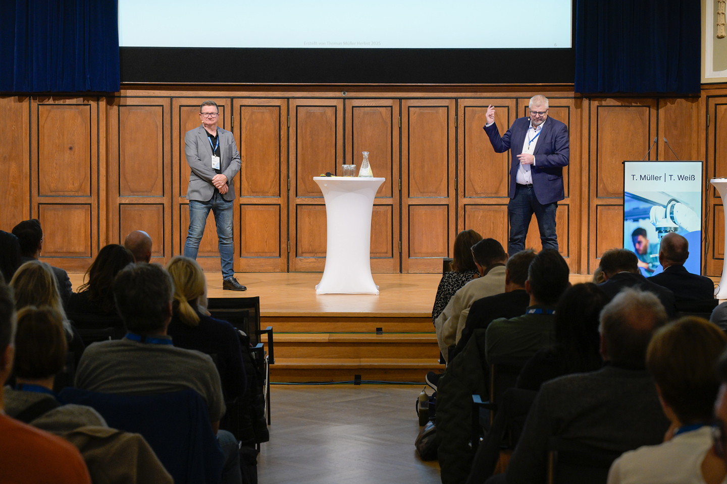 Zwei Männer stehen auf einer Bühne. Zwischen ihnen steht ein Stehtisch